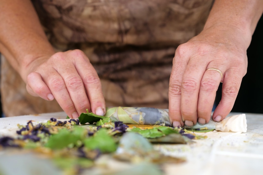 a woman's hands roll up a piece of silk that's been scattered with leaves and plants