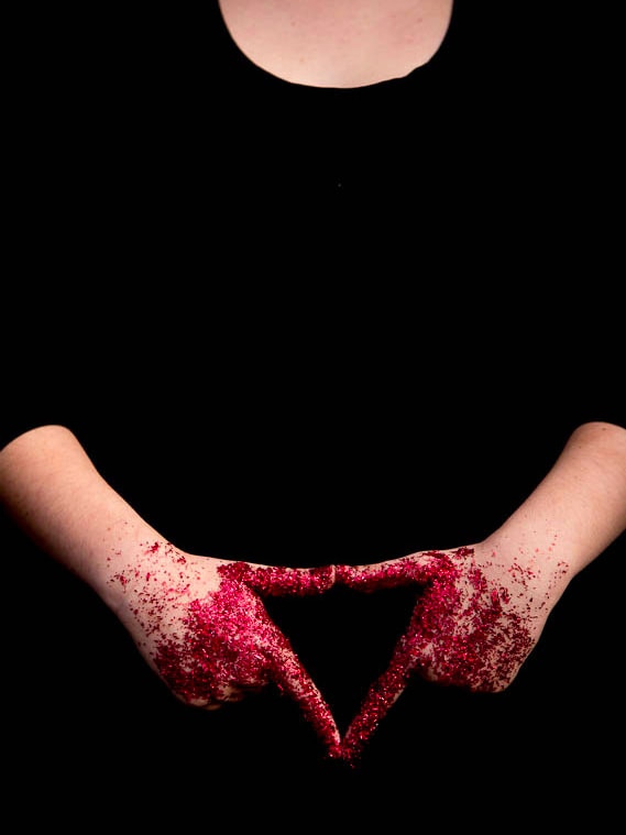 Hands covered in red paint form a triangle in front of a woman wearing a black dress.