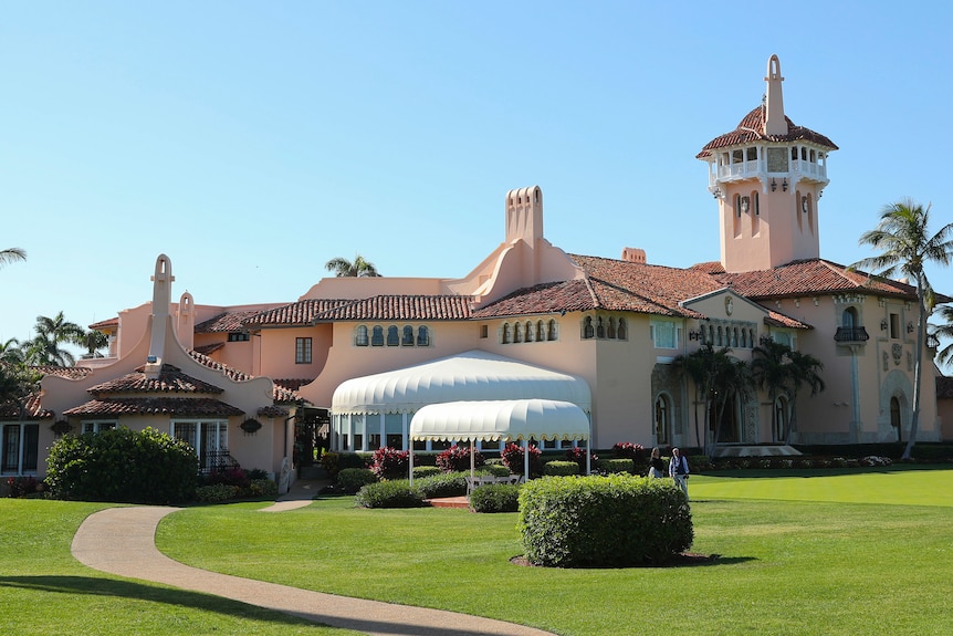 A pinkish Mediterranean-style villa sits on a manicured green lawn surrounded by palm trees under a bright blue sky.