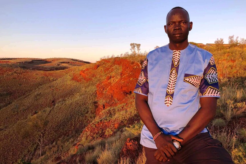 Philip Lako stands looking at the camera, with beautiful golden-hour red dirt hills and scrubland in the background.