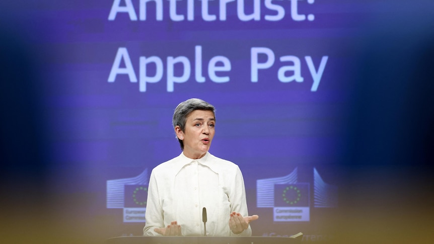 A woman stands at a microphone in front of a background with the words "Antitrust. Apple Pay"