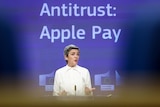 A woman stands at a microphone in front of a background with the words "Antitrust. Apple Pay"