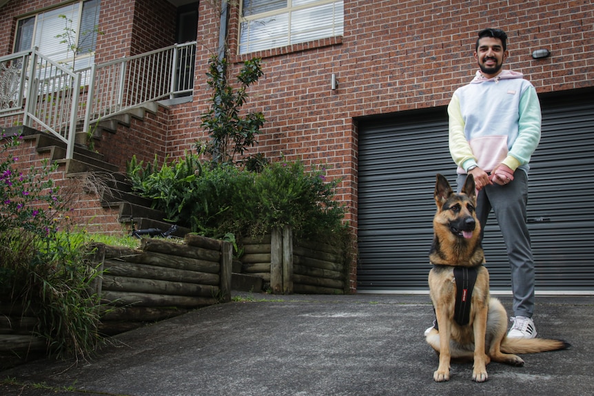 A man stands on stairs leading to his brick home with a German Shepherd dog at his feet.