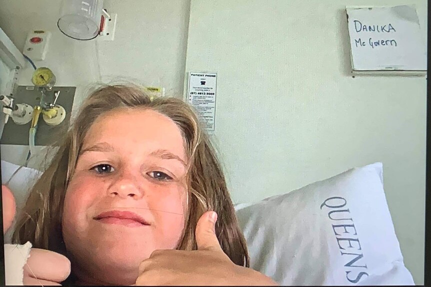 A young girl sits in a hospital bed and gives the thumbs up. 