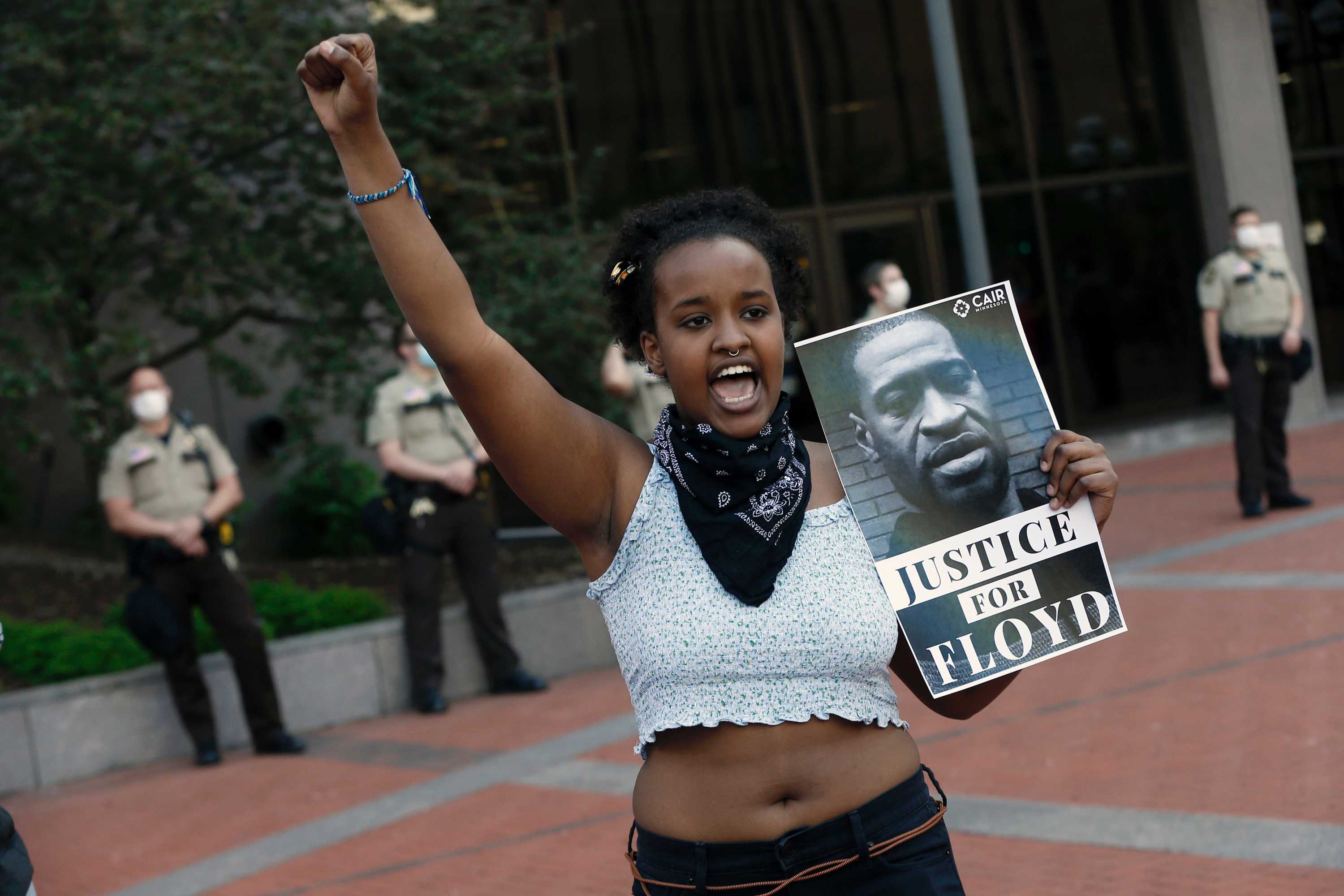 A young woman in white cut-off top raises her arm chants in street, holding Justice for Floyd sign.