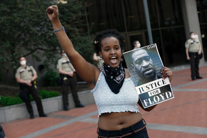 A young woman in white cut-off top raises her arm chants in street, holding Justice for Floyd sign.