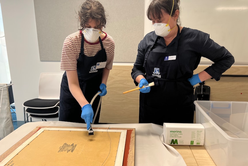 Two masked women clean a painting that has been damaged with mud.
