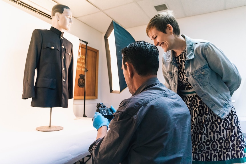 A man holds a camera and a woman looks at the camera screen over his shoulder, there's a uniform on a mannequin in front.