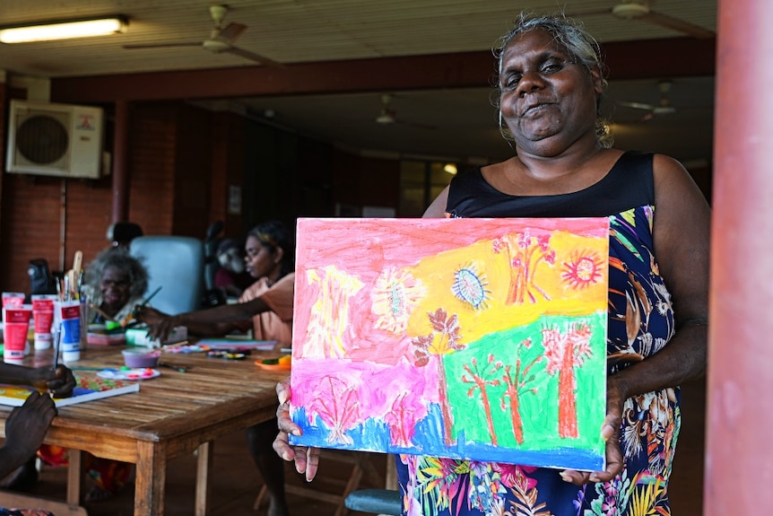 An elderly Aboriginal woman holds a colourful painting 