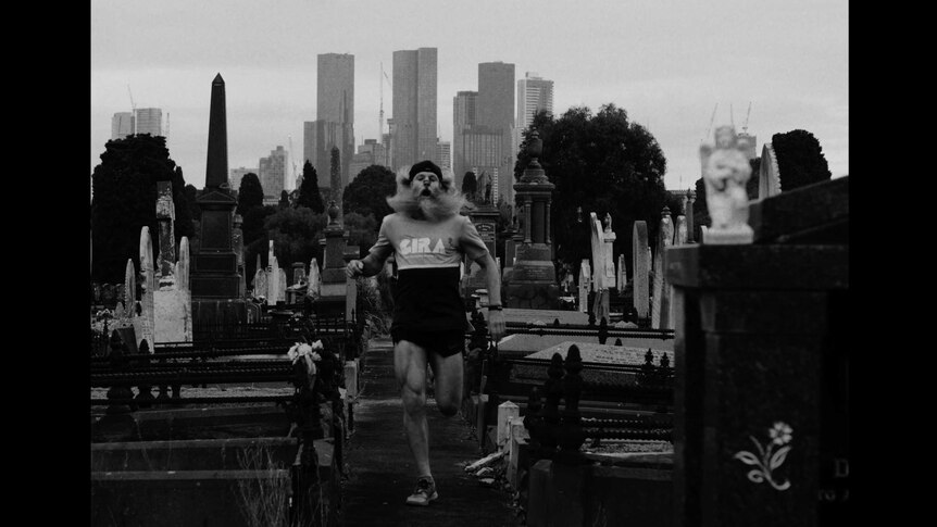 A man with long hair and a long beard is running through a graveyard, with the Melbourne skyline in the background