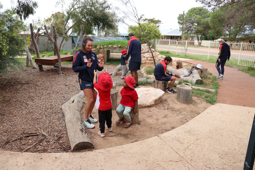 An AFLW player with long brown hair plays with some kids in a school playground.