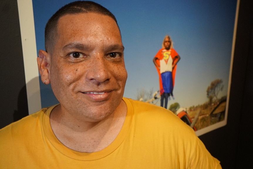 an aboriginal man wearing a yellow shirt in front of his artwork