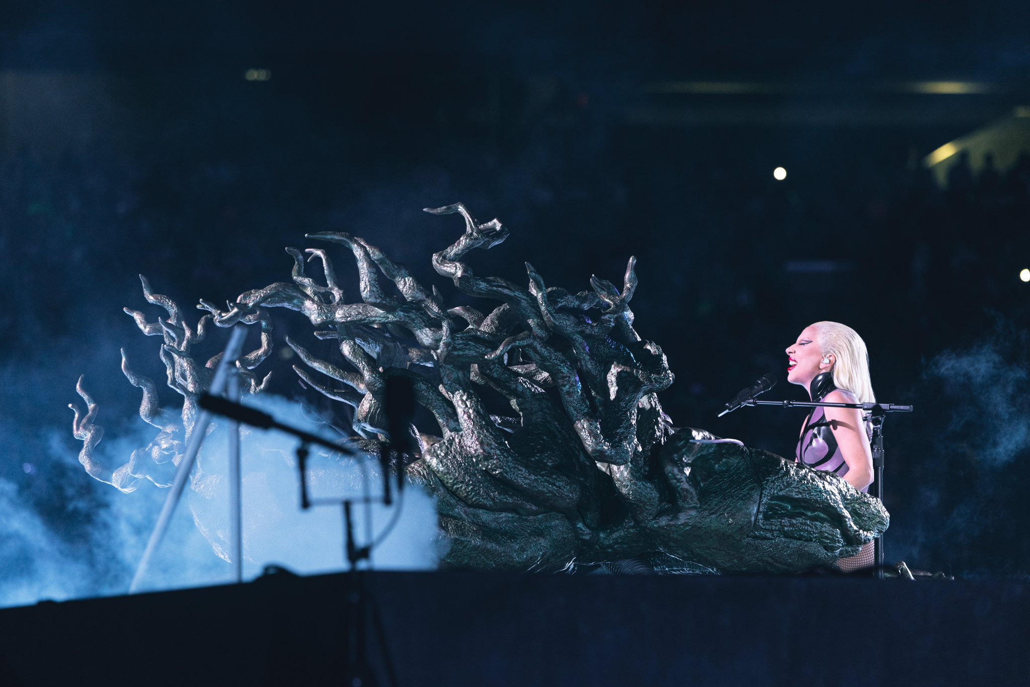 Lady Gaga sits at a piano made to look like a leafless tree.