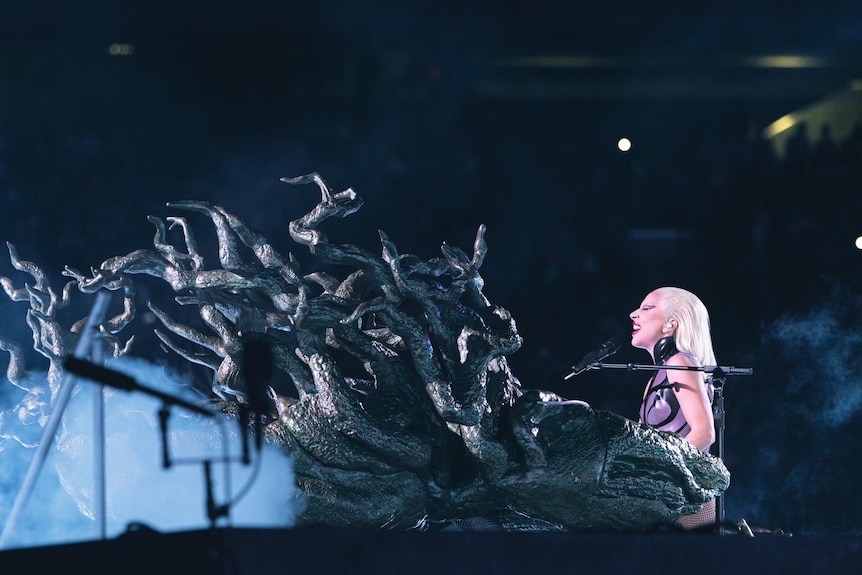 Lady Gaga sits at a piano made to look like a leafless tree.