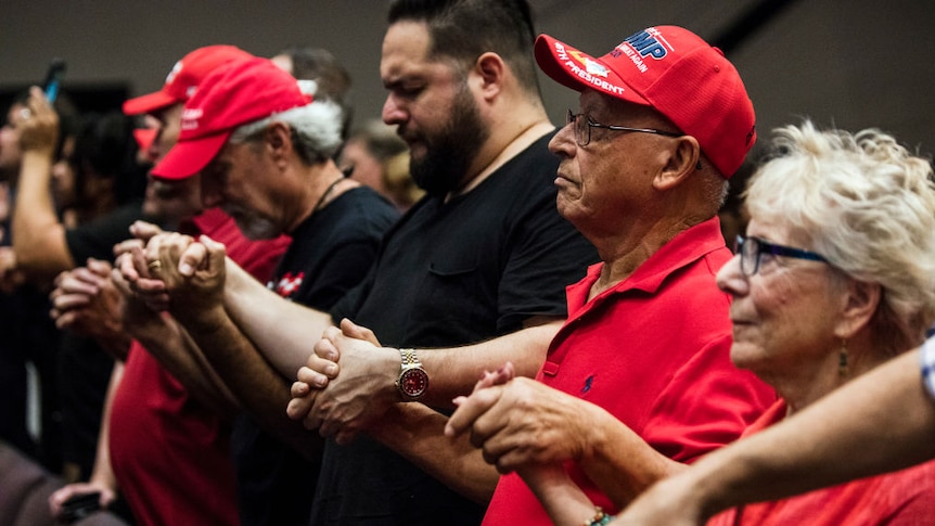 caucasian supporters dressed in red, some wearing Trump hats linking hands together