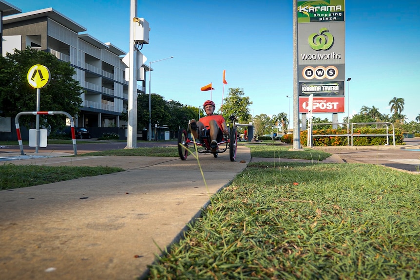 Man lying on a recumbent tricycle passing a bus stop and suburban shopping centre.