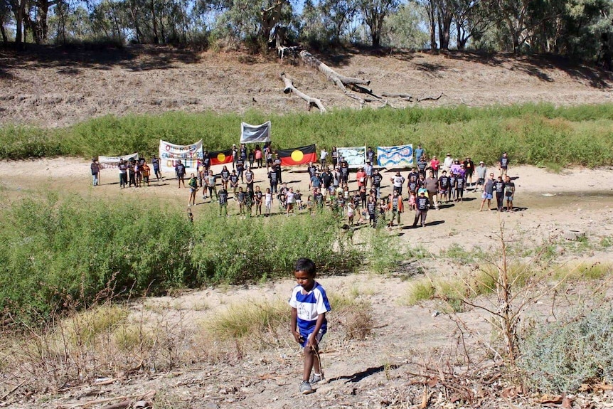 At least 80 people stand in a giant dry river bed holding banners and aboriginal flags, an aboriginal boy walks in foreground