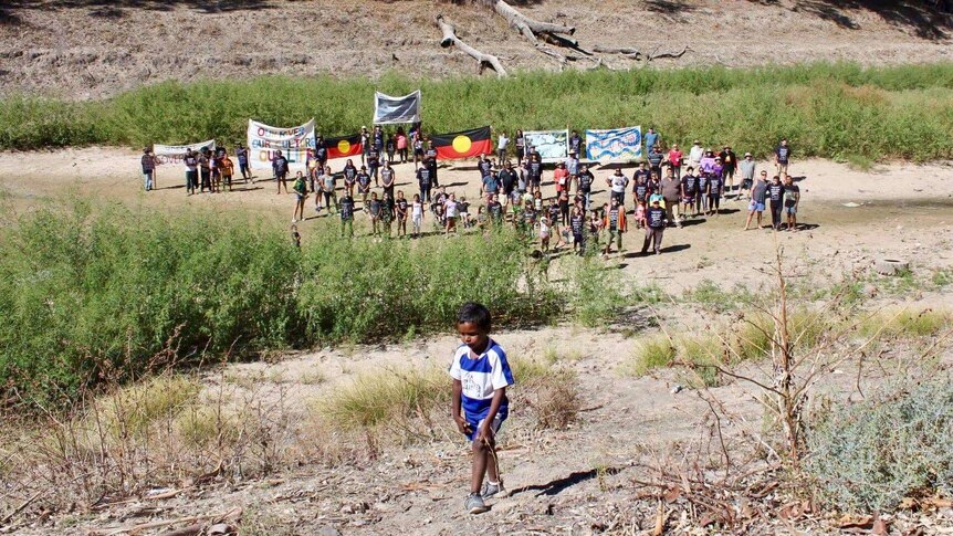 At least 80 people stand in a giant dry river bed holding banners and aboriginal flags, an aboriginal boy walks in foreground