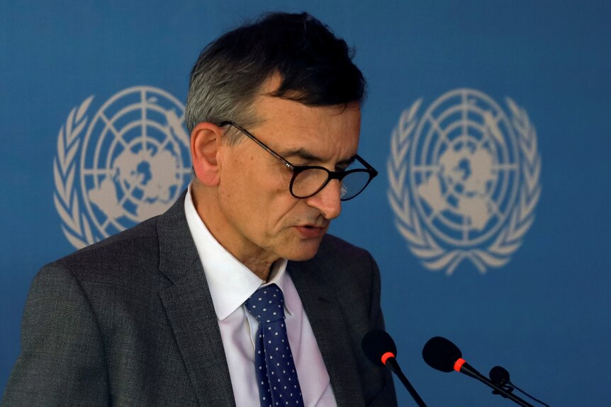 A man in glasses and a suit speaks into a mic in front of the UN logo. 