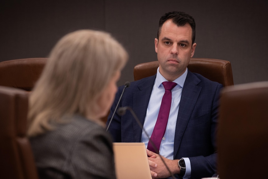 Man in a suit looks serious while looking at a pale-haired woman across the table