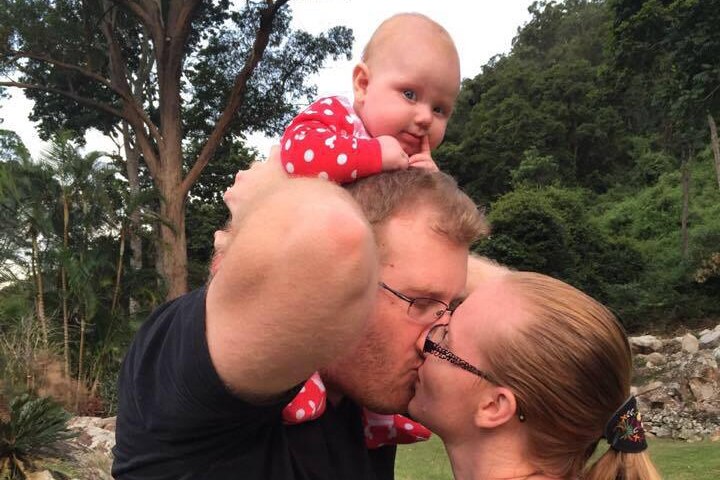 A woman kisses her husband, who has a baby on his shoulders.