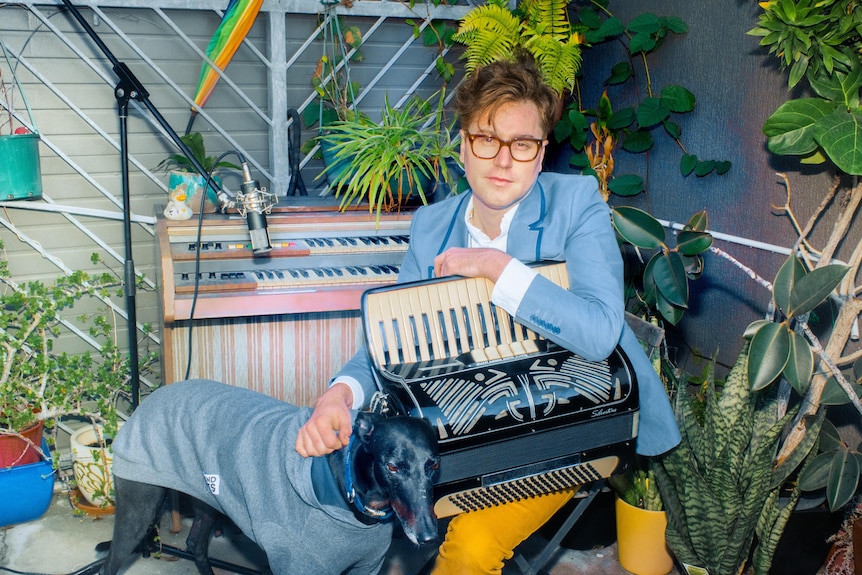 A man in a blue suit sits on a piano stool in front of a piano in an outdoor garden with a dog.