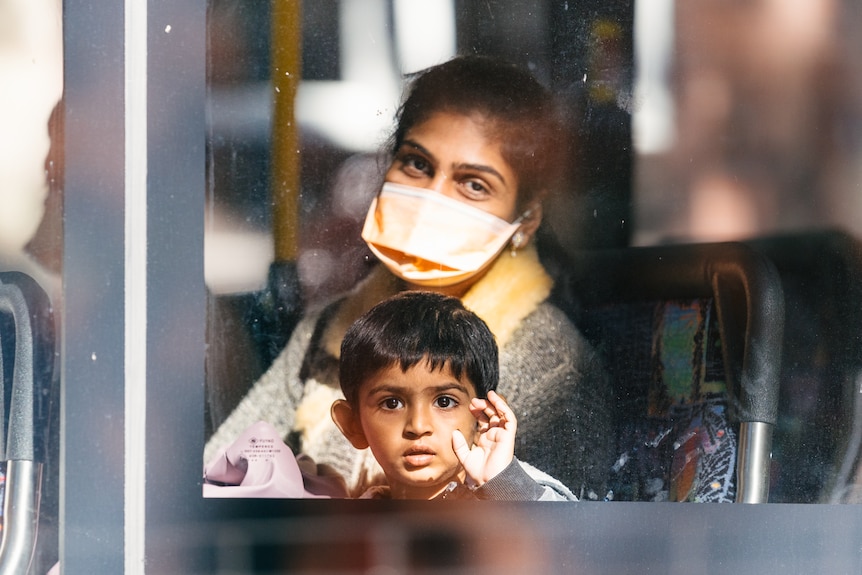 A woman wearing a face mask and a waving child look out of the window of a bus.