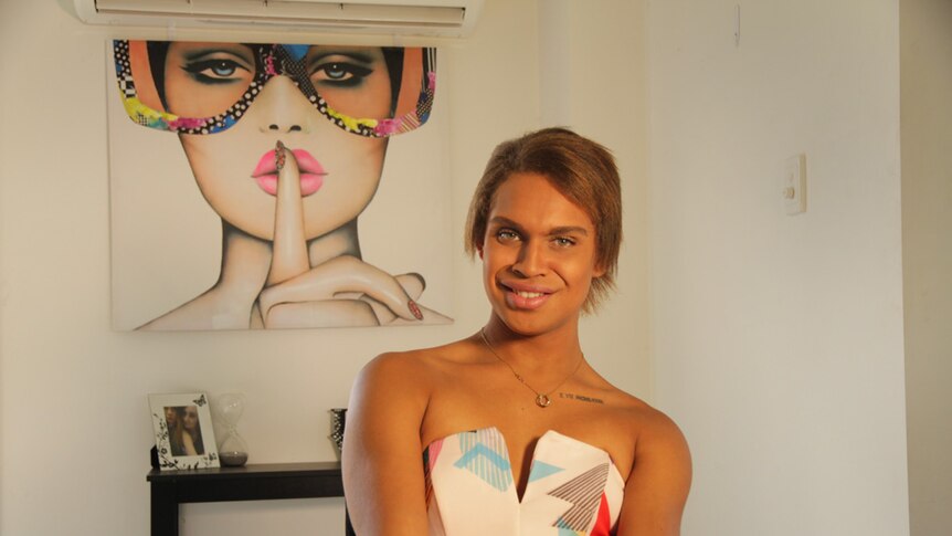A transgender Indigenous woman wearing a strapless dress, sitting in front of a painting.
