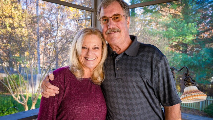 A man with a moustache wraps his arm around a shorter blonde woman while standing on a porch 