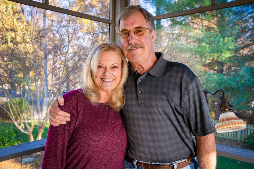 A man with a moustache wraps his arm around a shorter blonde woman while standing on a porch 