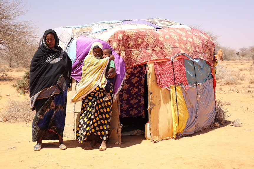 Two women, one holding a baby, stand in front of a tent.