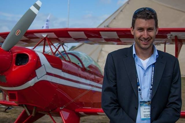 Ido Segev smiles as he stands next to a small red airplane with white stripes outside a large tent in a field.