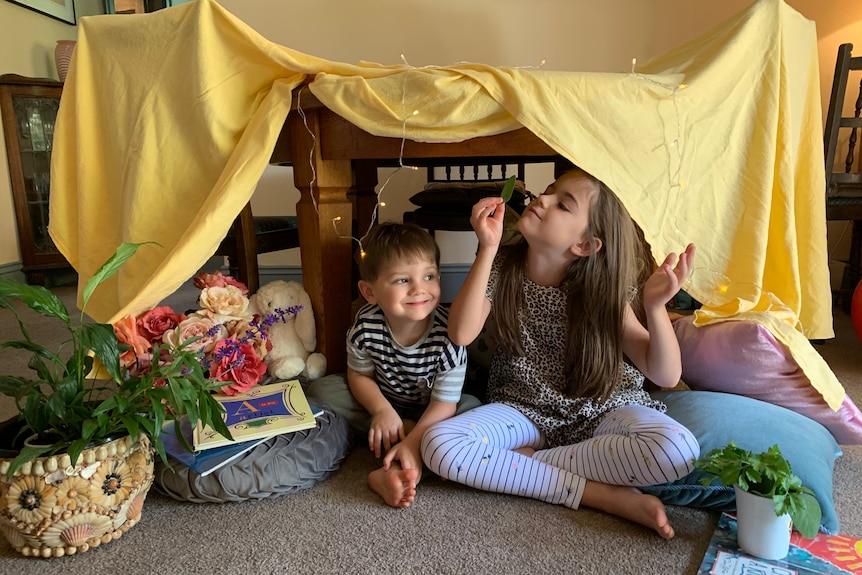 Two children sit underneath a table with a yellow cloth thrown over it, like a homemade cubby house.