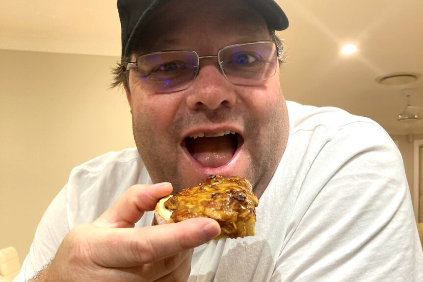 A man in his 40s wearing a baseball cap and glasses eats toast topped with melted cheese and bacon. 