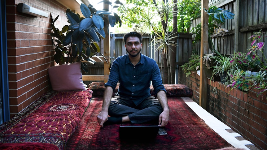 A man with dark hard and trimmed beard sits cross-legged in an outdoor courtyard, surrounded by plants and wooden fence 