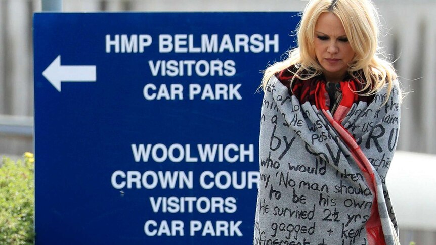 A blonde woman wrapped in a blanket with protest words written on it stands in front of a sign to a prison.