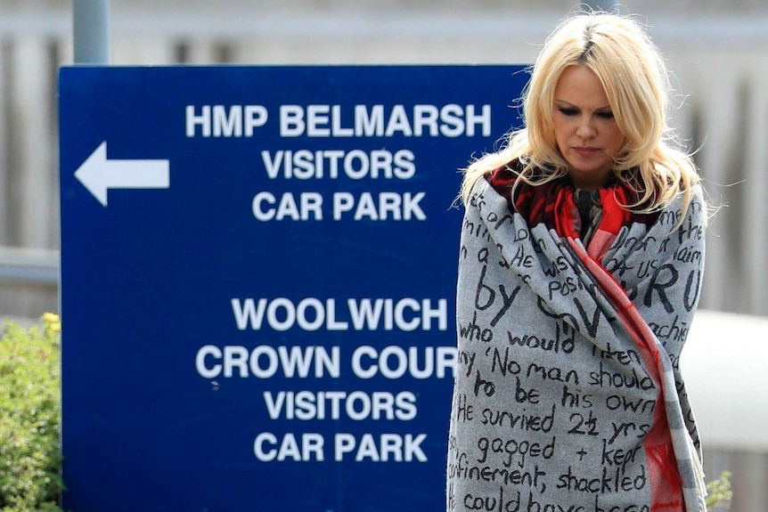 A blonde woman wrapped in a blanket with protest words written on it stands in front of a sign to a prison.