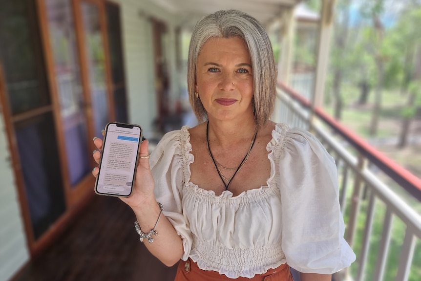 A mid shot of a woman in the foreground holding a mobile phone, with a blurred verandah in the background