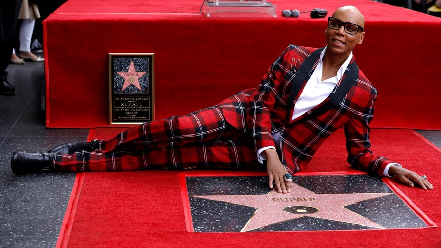 RuPaul in a red and black tartan suits lays next to a Hollywood star bearing his name