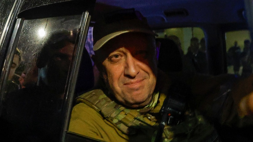 Yevgeny Prigozhin's face is illuminated by a streetlight as he partially smiles through the window of a car, rolled down