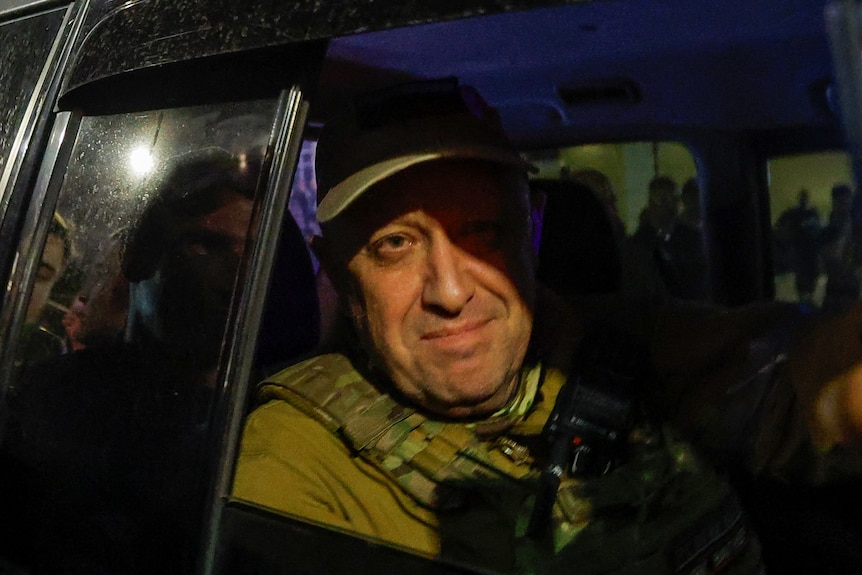 Yevgeny Prigozhin's face is illuminated by a streetlight as he partially smiles through the window of a car, rolled down