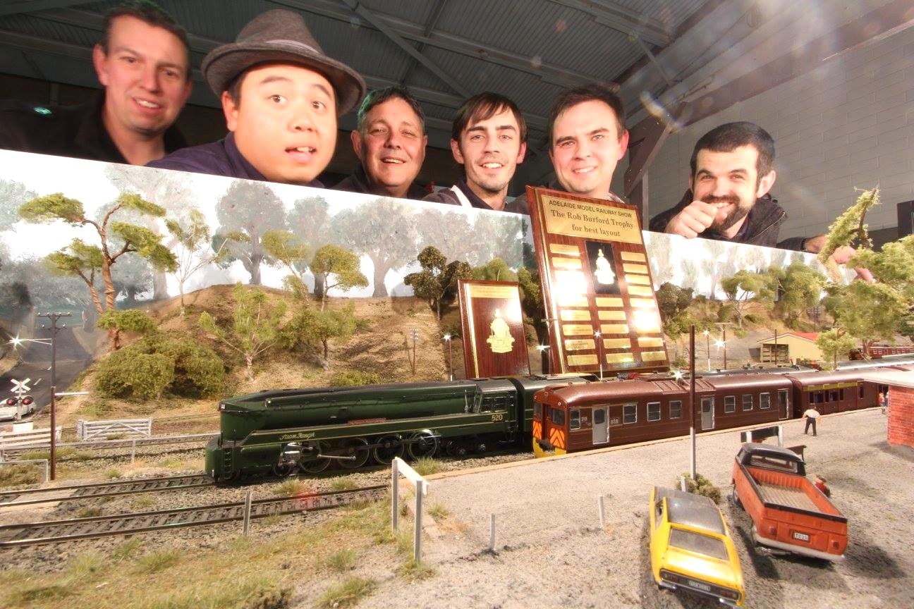 A group of six men pose behind a model railway scene and two plaques