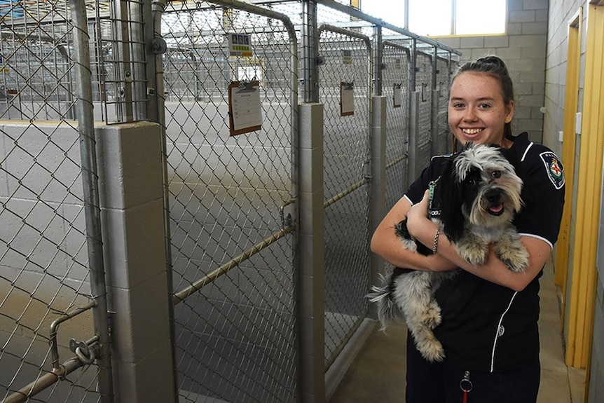 a girl smiles, while holding a dog near a group of silver cages. 