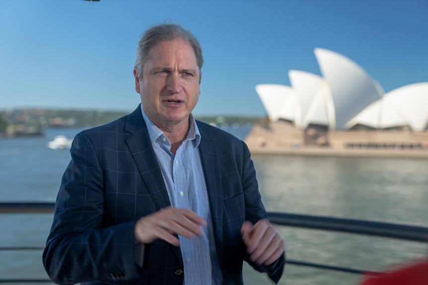 A man in a jacket stands at Circular Quay in front of the Sydney Opera House