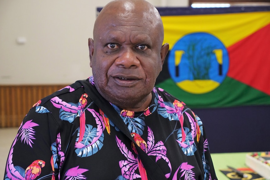 Kerry Warkill, dark skin, brown eyes, bald, wearing bright patterned t-shirt looks at the camera.