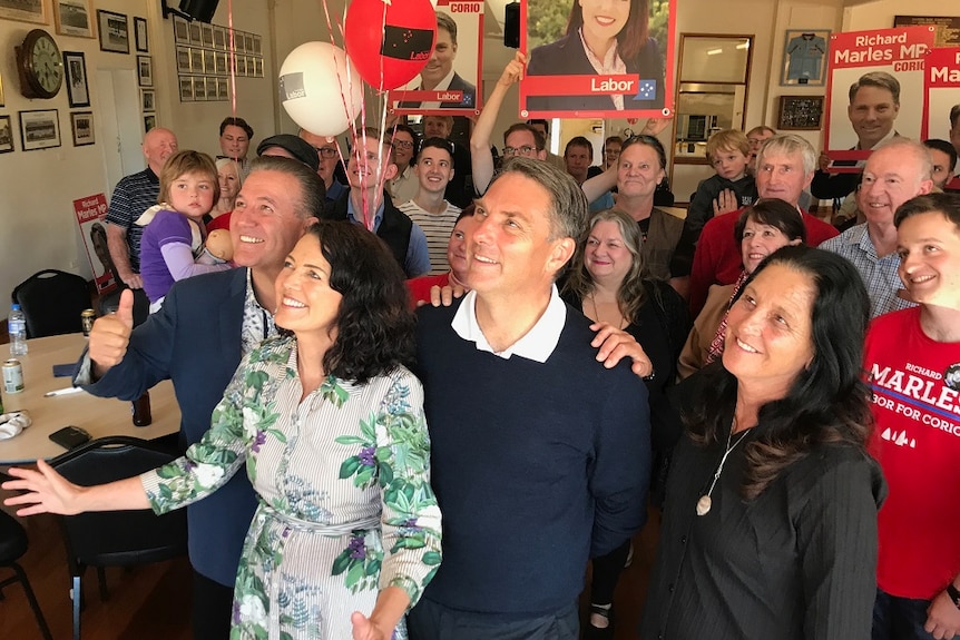 Libby Coker and Richard Marles stand surrounded by Labor supporters in a hall as they smile and look up into the light.