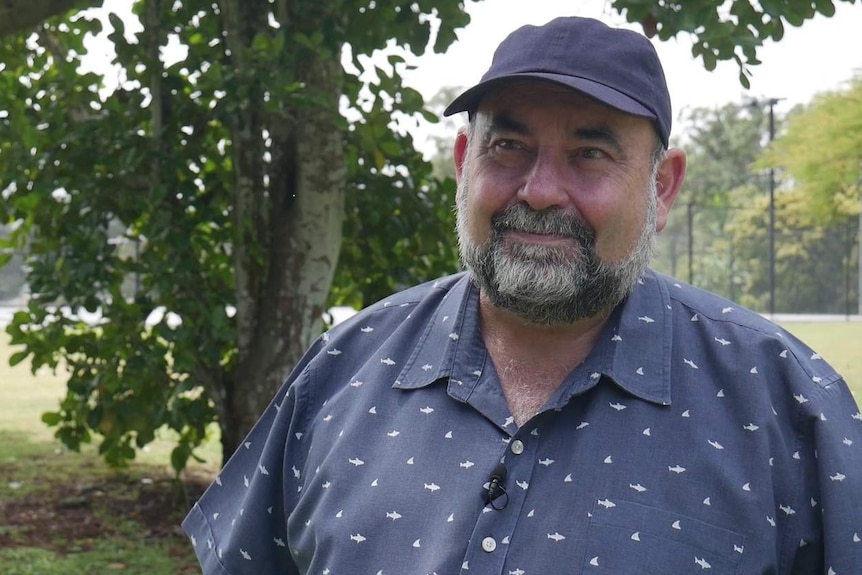 A bearded middle-aged man in a dark blue shirt and matching cap looks ahead in front of lush trees.