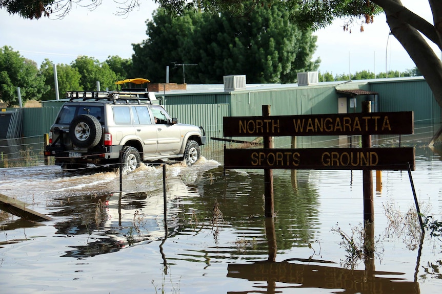 A four wheel drive drives through floodwaters near a sign that says 'North Wangaratta Sports Ground'.