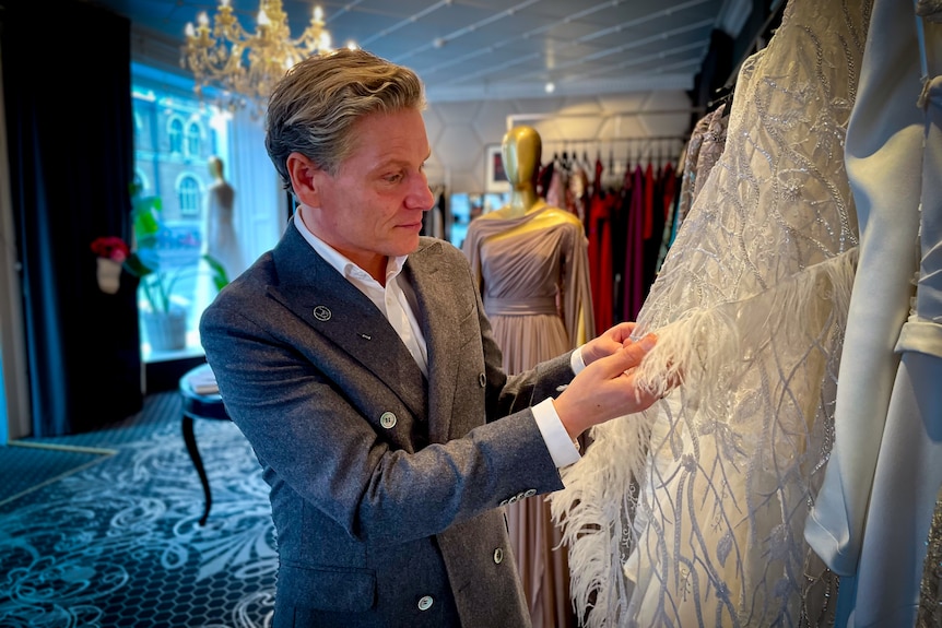 A man in a blazer inspects the hem of white beaded and feather-trimmed gown