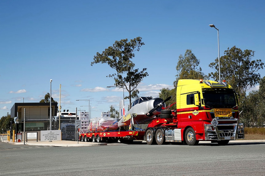 The body of a grey aircraft sits on the back of a red and yellow semi-trailer.